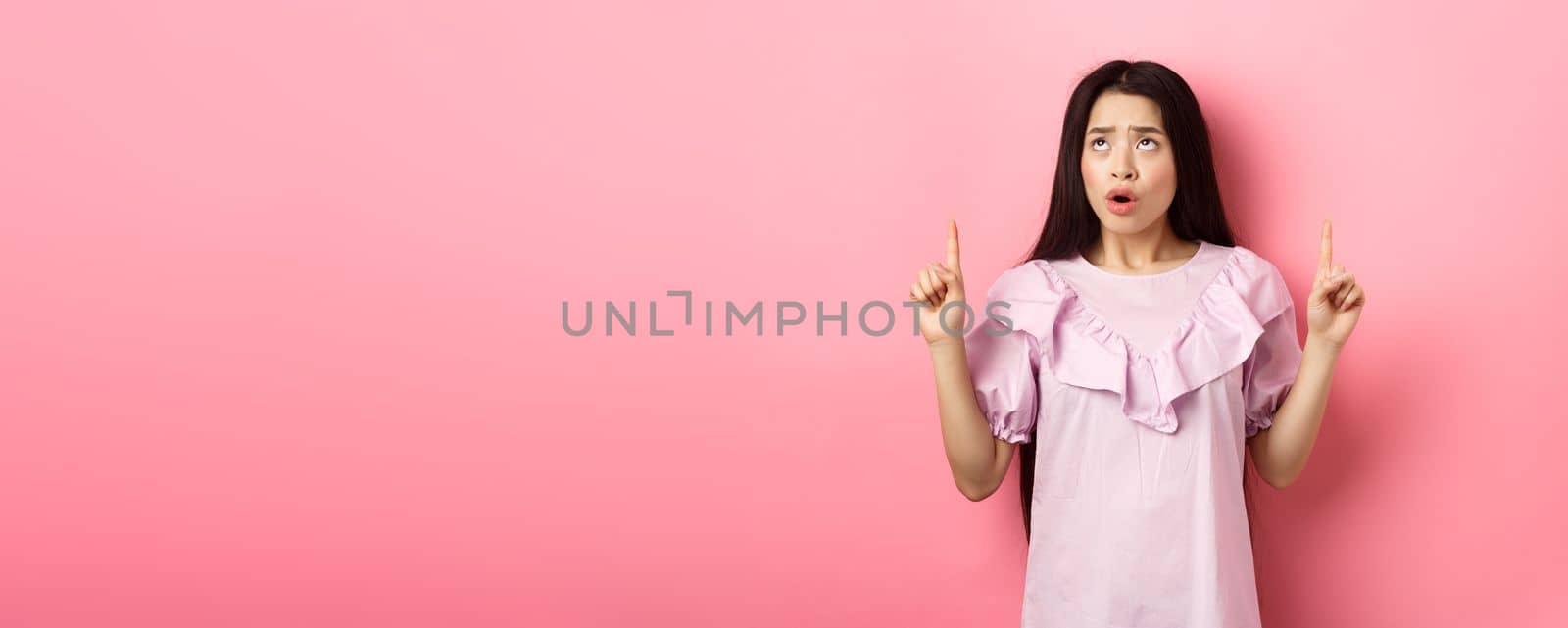 Concerned and sad asian girl frowning, looking and pointing fingers up at bad news, standing against pink background by Benzoix