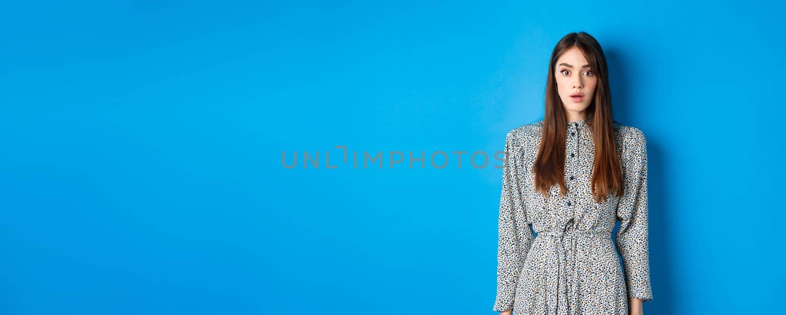 Shocked caucasian woman with long hair, wearing dress, gasping and standing in stupid against blue background, stare with disbelief by Benzoix