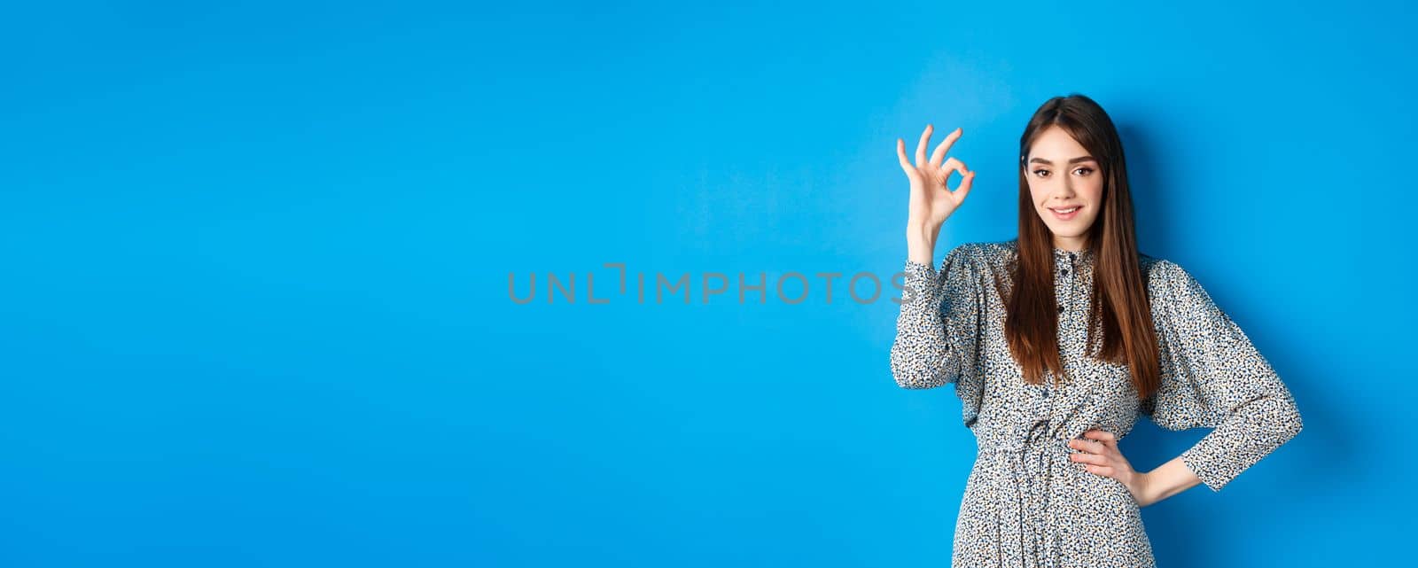 Confident young woman in dress showing okay sign and smiling, no problem gesture, assure everything good, standing on blue background by Benzoix