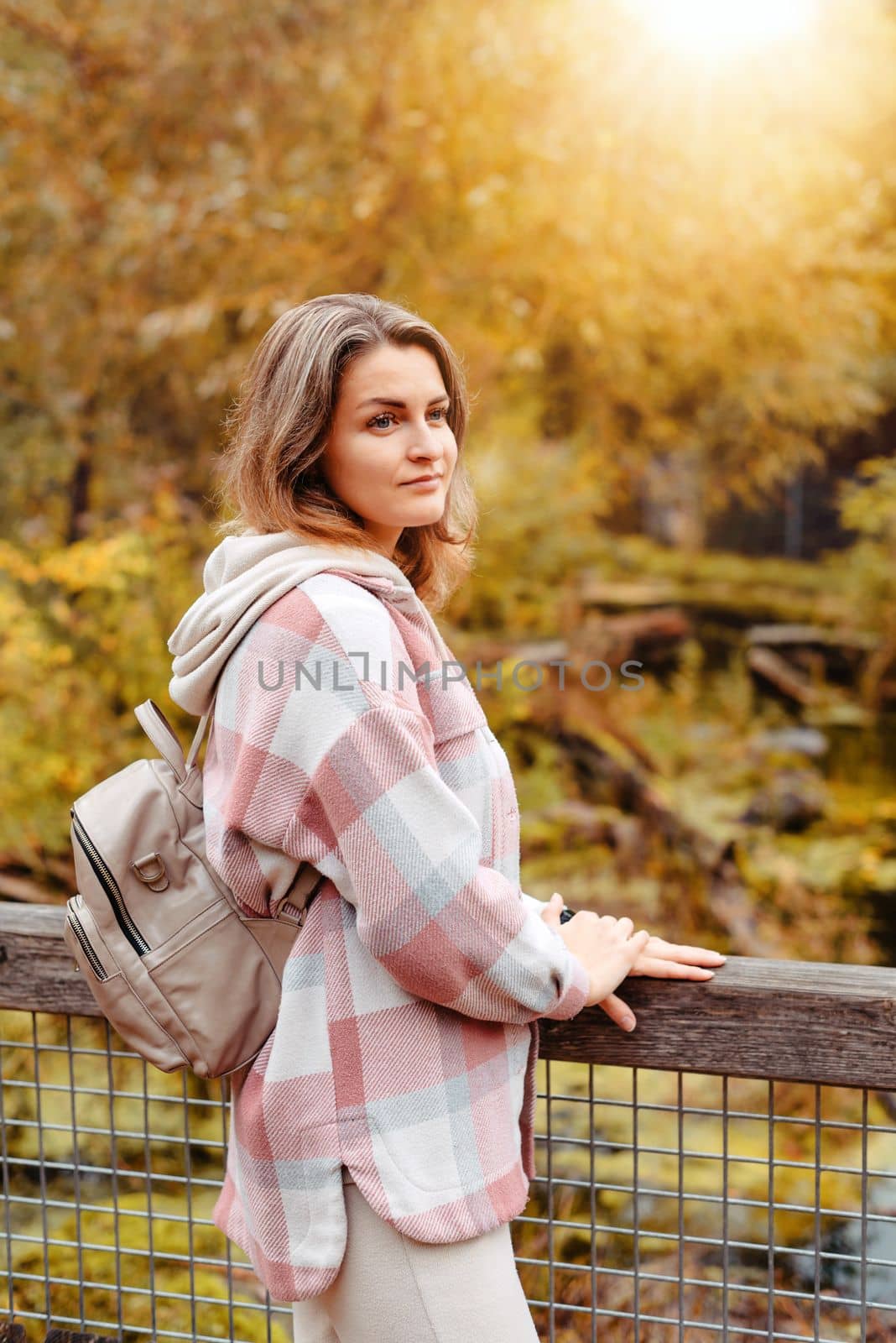 Portrait of cute young woman in casual wear in autumn, standing on bridge against background of an autumn Park and river. Pretty female walking in Park in golden fall. Copy space. smiling girl in the park standing on wooden bridge and looking at the camera in autumn season