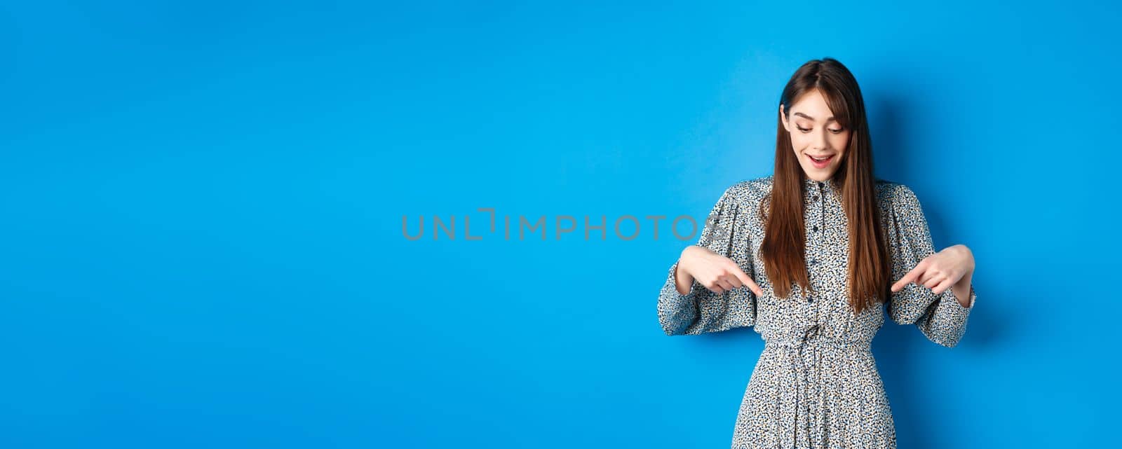 Dreamy happy woman pointing fingers down and looking wondered at promotion, showing awesome news, standing in dress on blue background by Benzoix