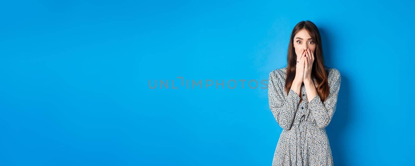 Shocked european girl gasping and covering mouth with hands, witness something and look startled, standing worried on blue background by Benzoix