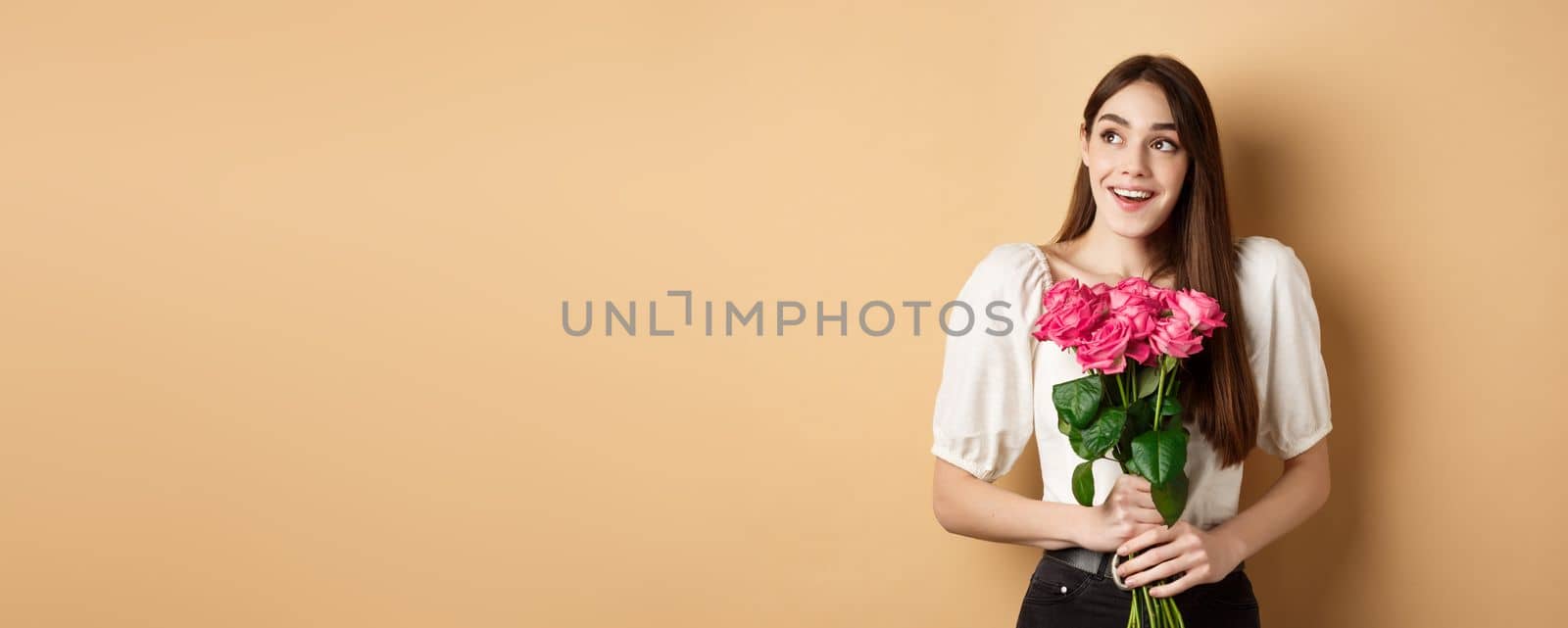 Happy valentines day. Dreamy smiling woman receive bouquet of pink roses and looking aside with love, standing on beige background by Benzoix