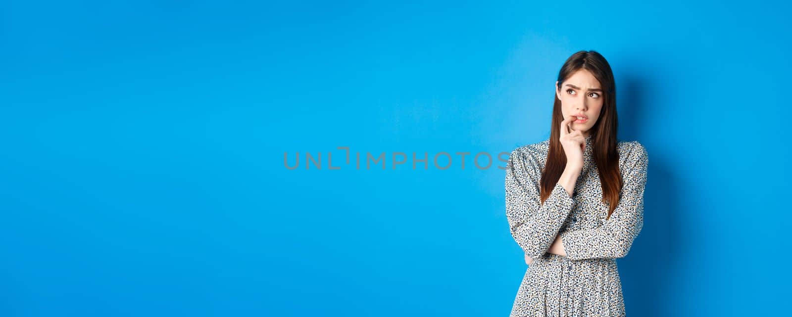 Troubled pensive girl looking aside at logo, biting fingernail and thinking, standing on blue background in long dress.