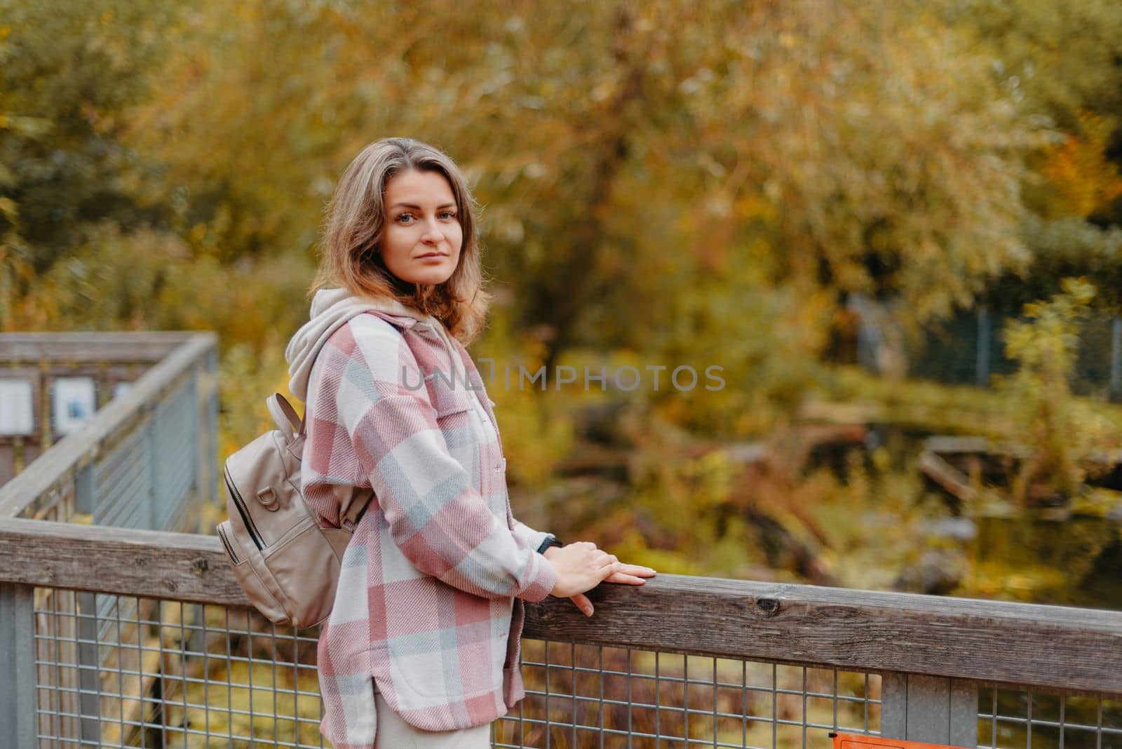 Portrait of cute young woman in casual wear in autumn, standing on bridge against background of an autumn Park and river. Pretty female walking in Park in golden fall. Copy space. smiling girl in the park standing on wooden bridge and looking at the camera in autumn season