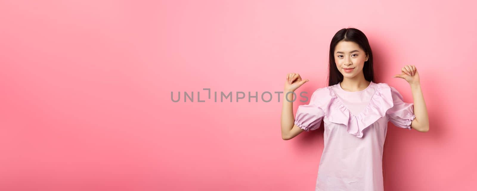 Confident asian girl smiling, pointing at herself, self-promoting personal achievements, standing in dress on pink background.
