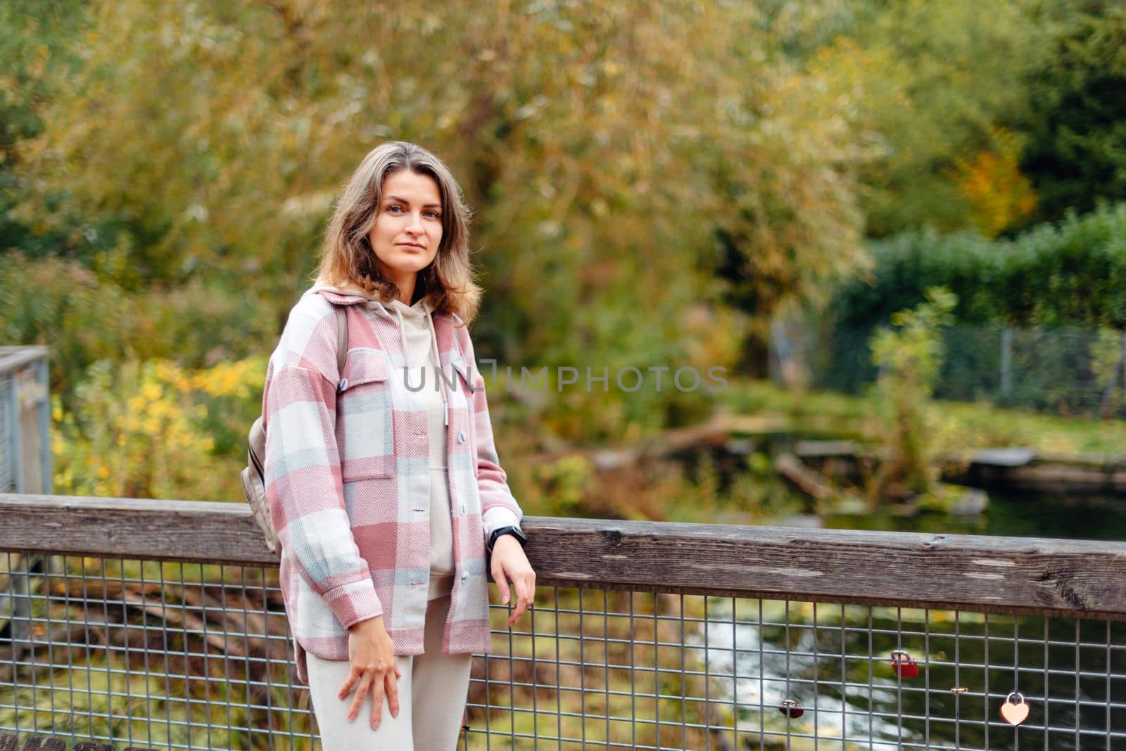 Portrait of cute young woman in casual wear in autumn, standing on bridge against background of an autumn Park and river. Pretty female walking in Park in golden fall. Copy space. smiling girl in the park standing on wooden bridge and looking at the camera in autumn season