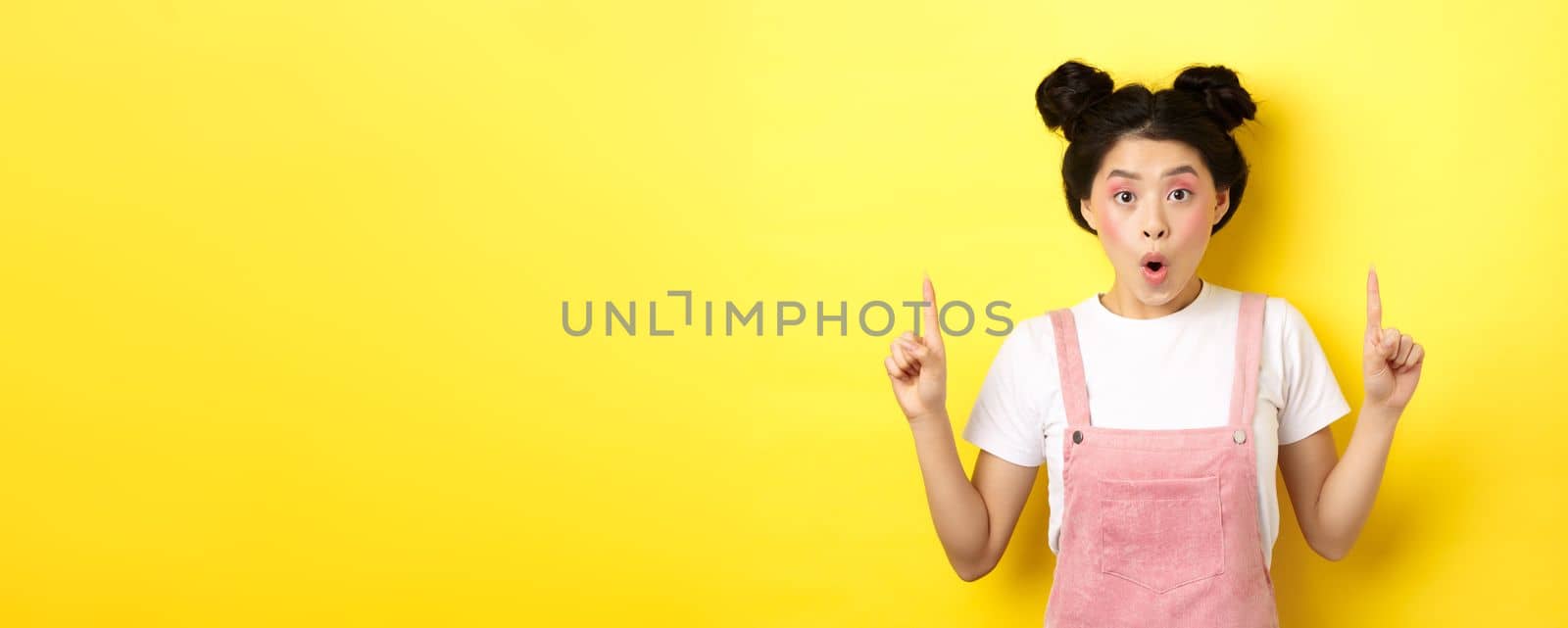 Excited asian female model with glamour makeup, pointing fingers up and say wow amazed, checking out promo deal, standing on yellow background.