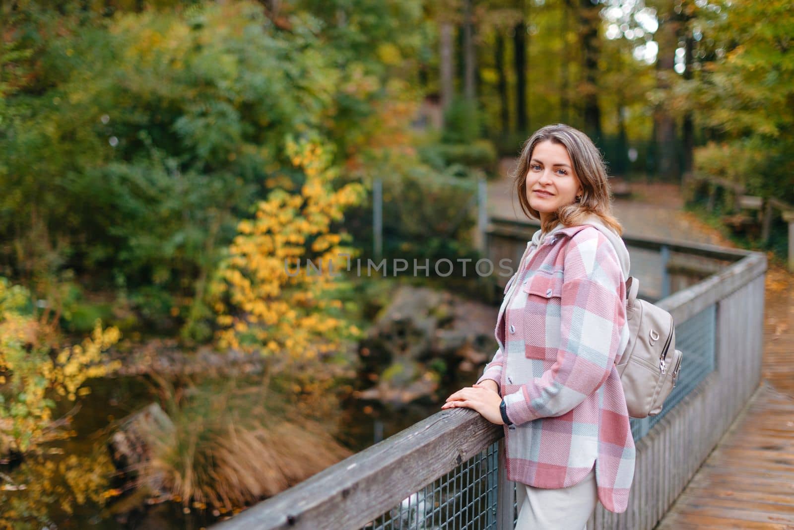 Portrait Of Cute Young Woman In Casual Wear In Autumn, Standing On Bridge Against Background Of An Autumn Park And River. Pretty Female Walking In Park In Golden Fall. Copy Space. Smiling Girl In The Park Standing On Wooden Bridge And Looking At The Camera In Autumn Season by Andrii_Ko