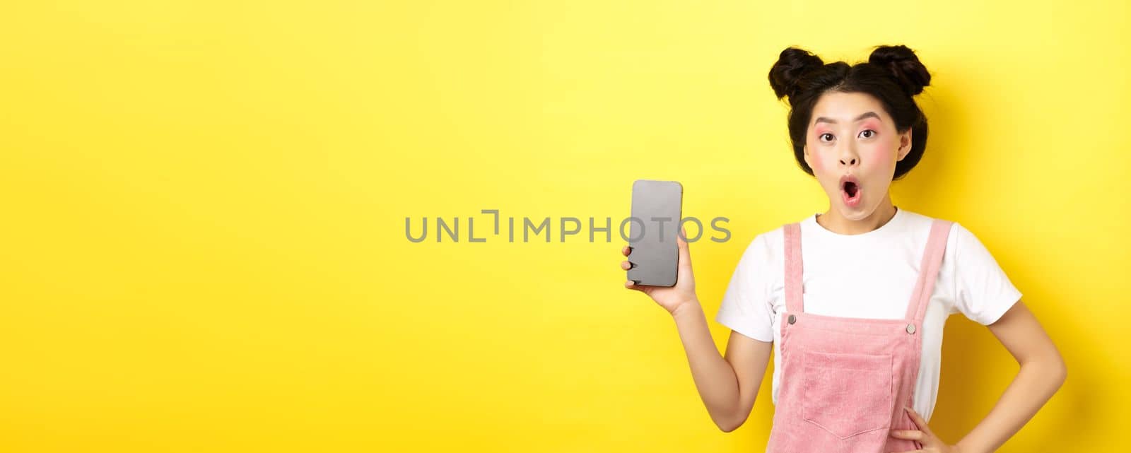 Online shopping concept. Excited stylish asian girl say wow, showing empty cell phone screen and look amazed, yellow background.