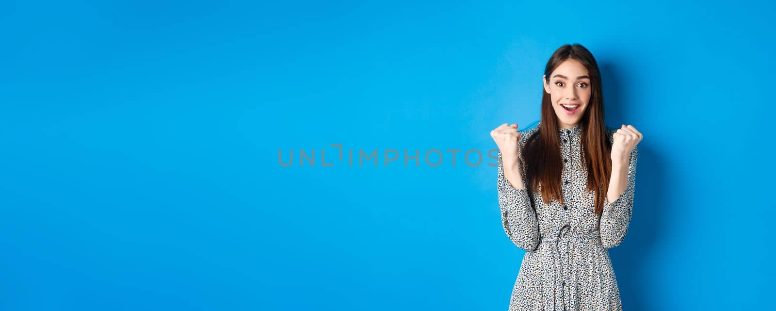 Excited woman looking relieved and hopeful, making fist pump and smiling, winning prize, celebrating achievement or victory, standing on blue background.