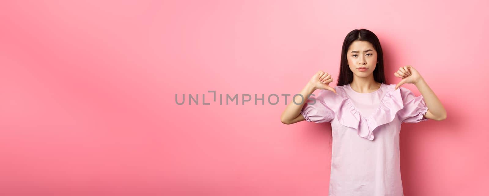 Skeptical and disappointed asian woman showing thumbs down with reluctant smirk, dislike product, show negative emotion, standing on pink background.