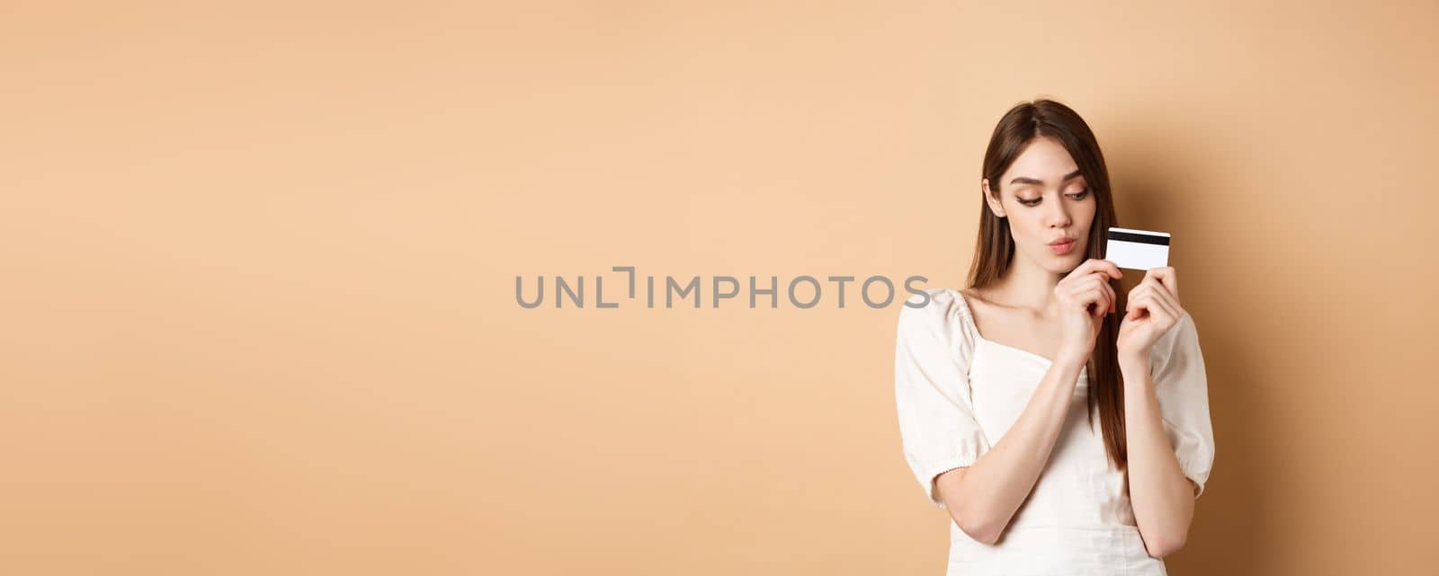 Cute young woman looking thoughtful at plastic credit card, thinking of shopping, standing on beige background.