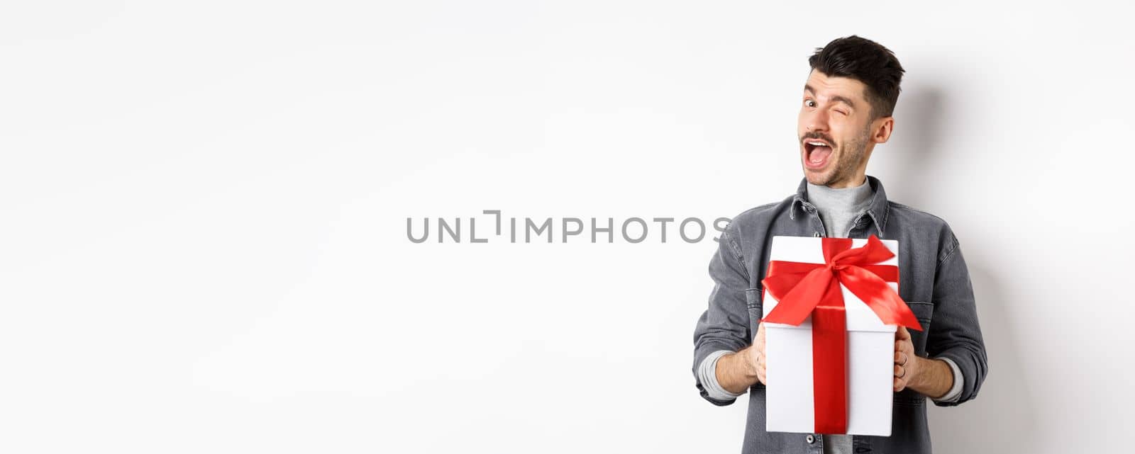 Funny young man holding valentines gift and winking at camera, making romantic surprise to lover, standing on white background.