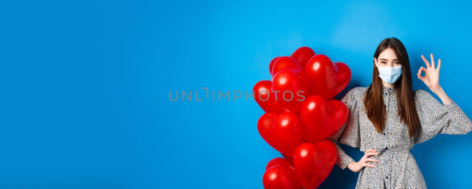 Covid-19, Valentines day and pandemic concept. Beautiful young woman in face mask and dress, showing OK sign in approval, standing near romantic red hearts balloons, blue background.