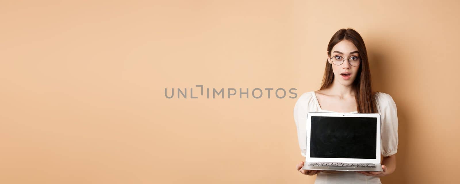 Surprised young woman in glasses showing laptop screen with amazed face, standing on beige background.