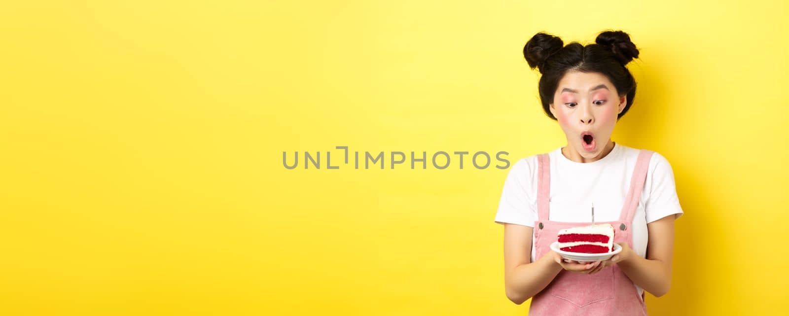 Cute asian birthday girl with bright makeup, blowing candle on cake, making wish, standing on yellow background.
