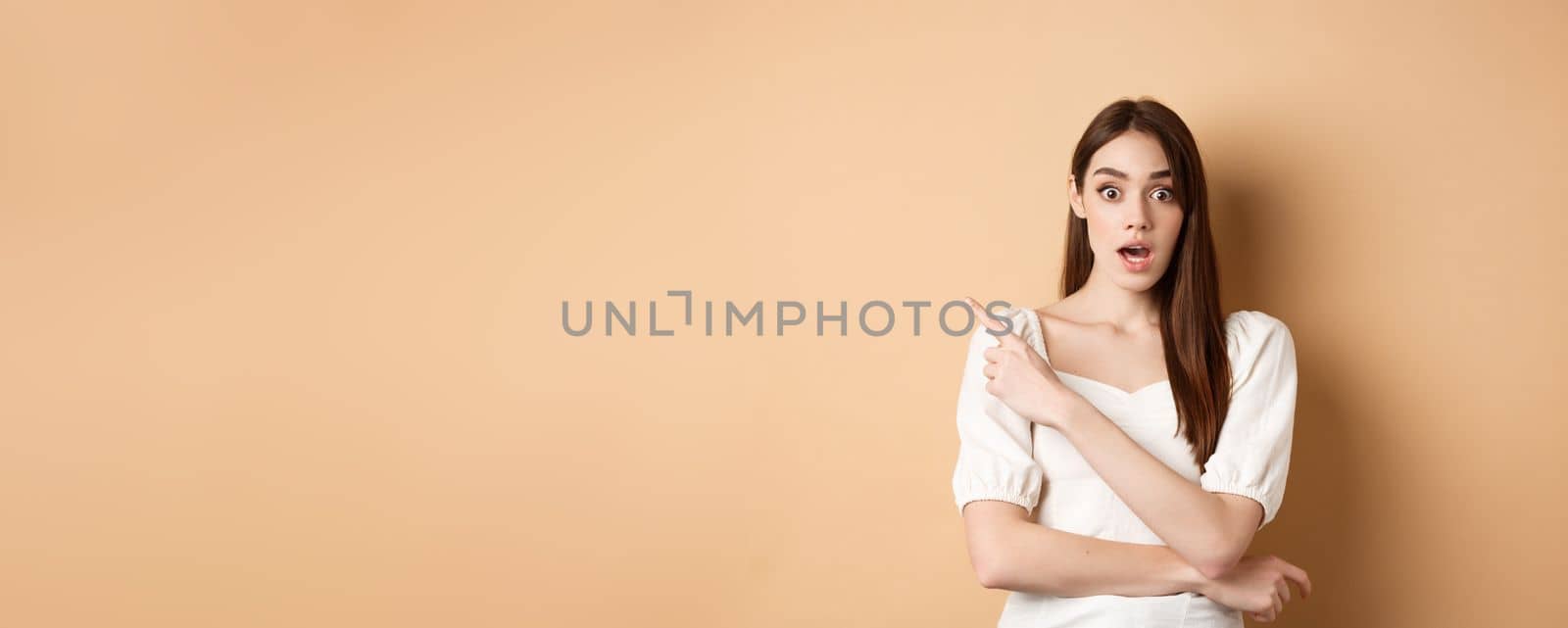 Surprised young girl say wow, pointing finger left at interesting banner, standing curious against beige background by Benzoix