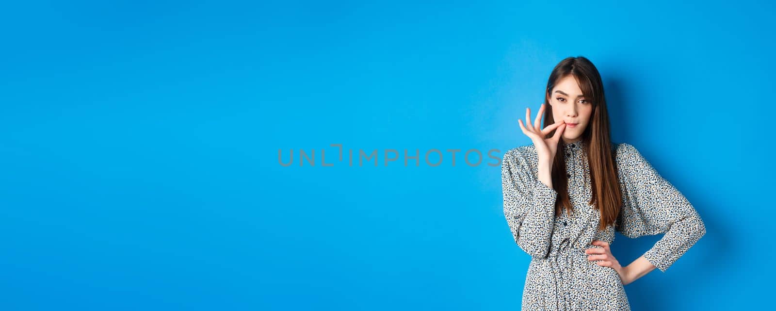 Serious woman promise to keep secret, seal lips, making zip gesture and looking at camera, standing in dress on blue background by Benzoix