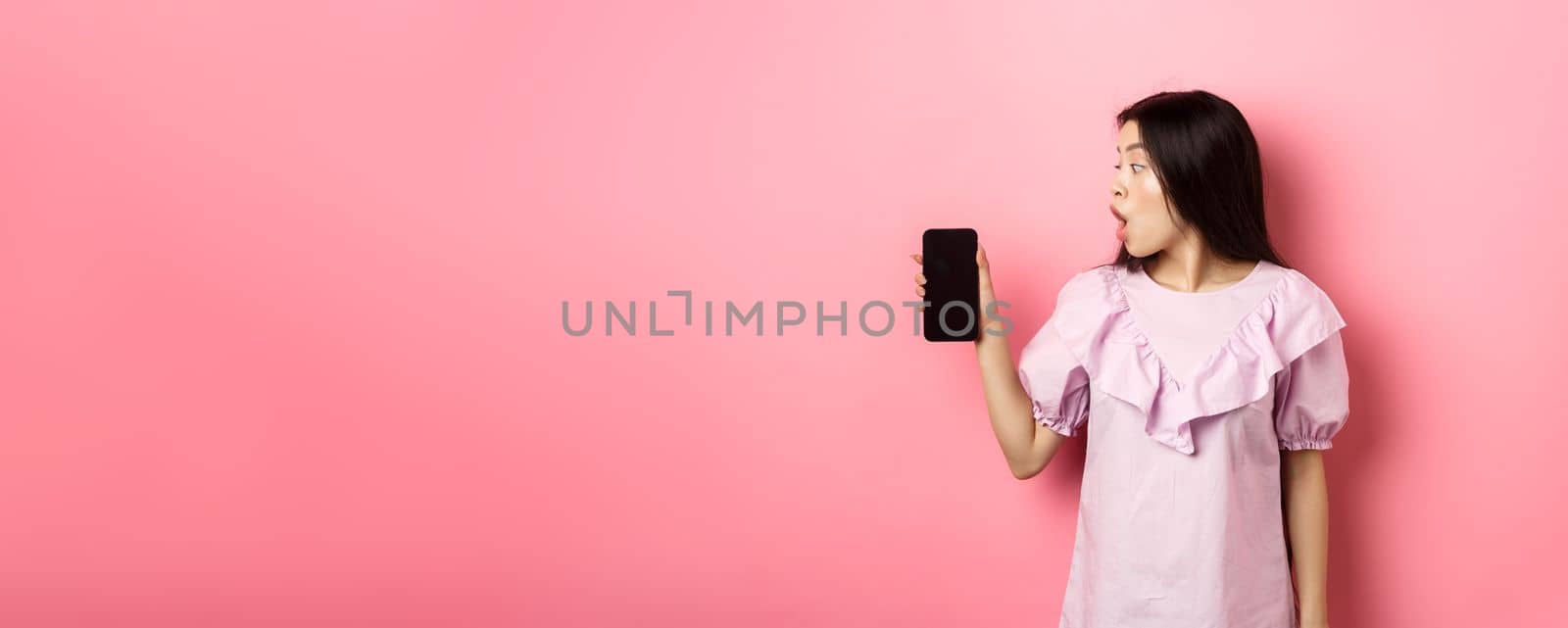 Wow look here. Excited asian girl look at smartphone screen with amazed face, checking out online offer, standing against pink background.