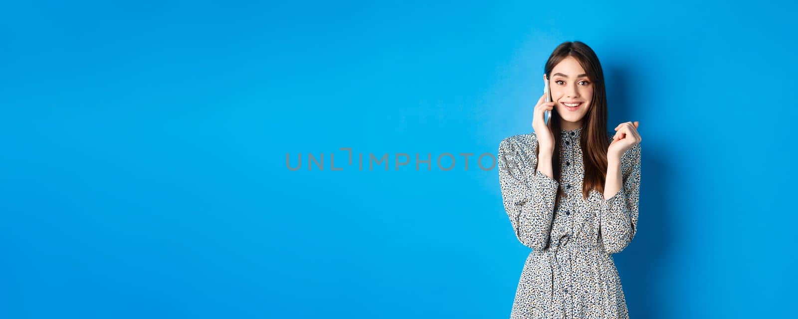 Excited woman receive good news on mobile phone, smiling happy, talking on smartphone, standing on blue background.