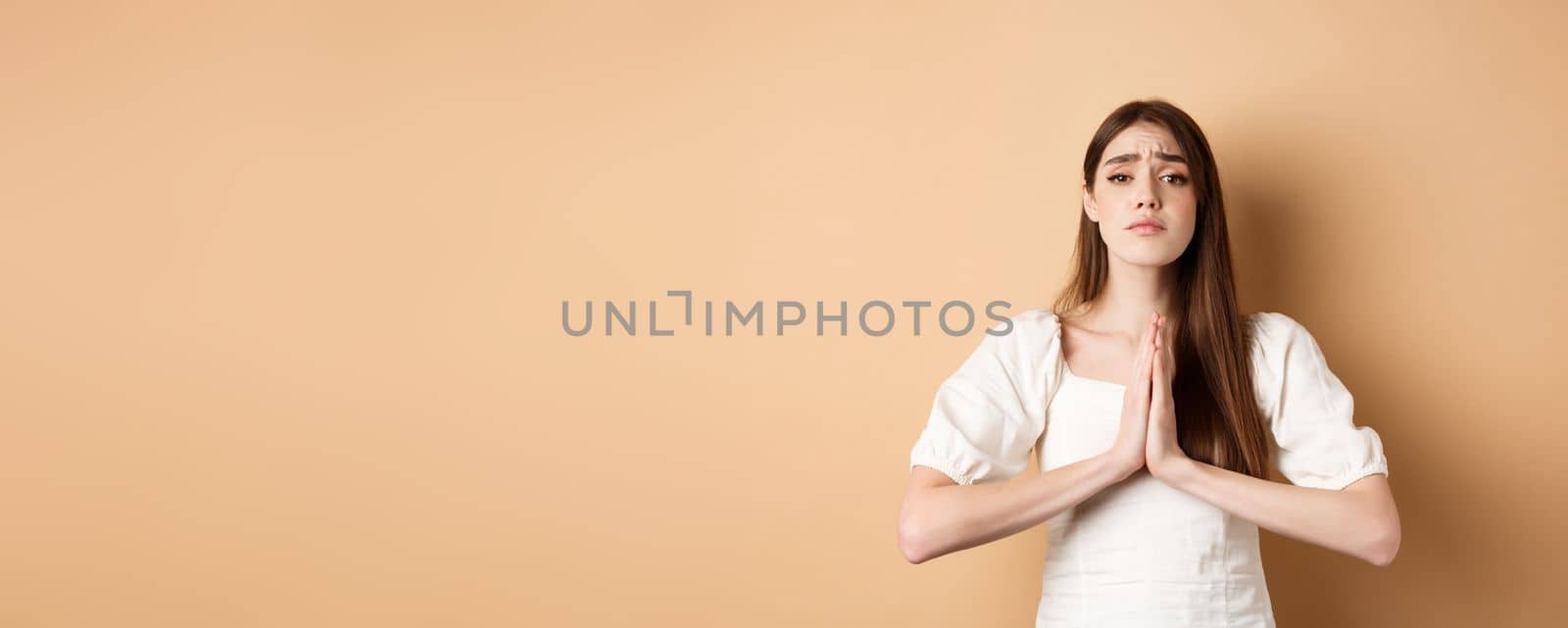 Please help. Desperate young woman begging you, holding hands in pleading gesture and looking sad at camera, asking for favour, beige background.