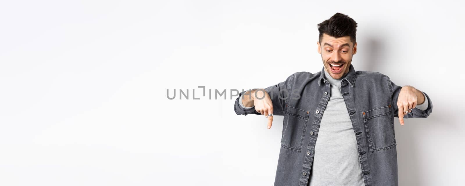 Excited european man looking and pointing fingers down, looking happy at advertisement, standing in denim jacket on white background by Benzoix