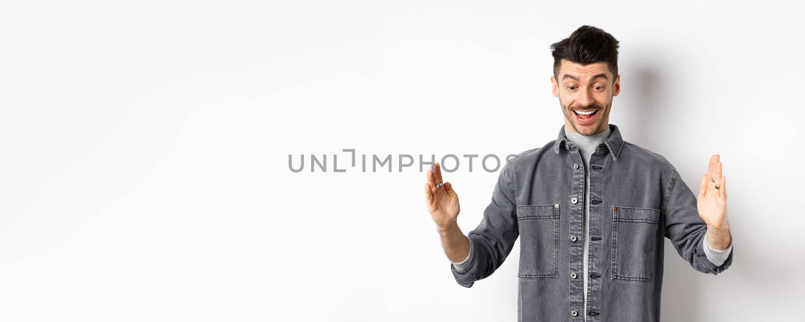 Excited caucasian man showing big size object, shaping large thing and smiling amazed, standing on white background by Benzoix