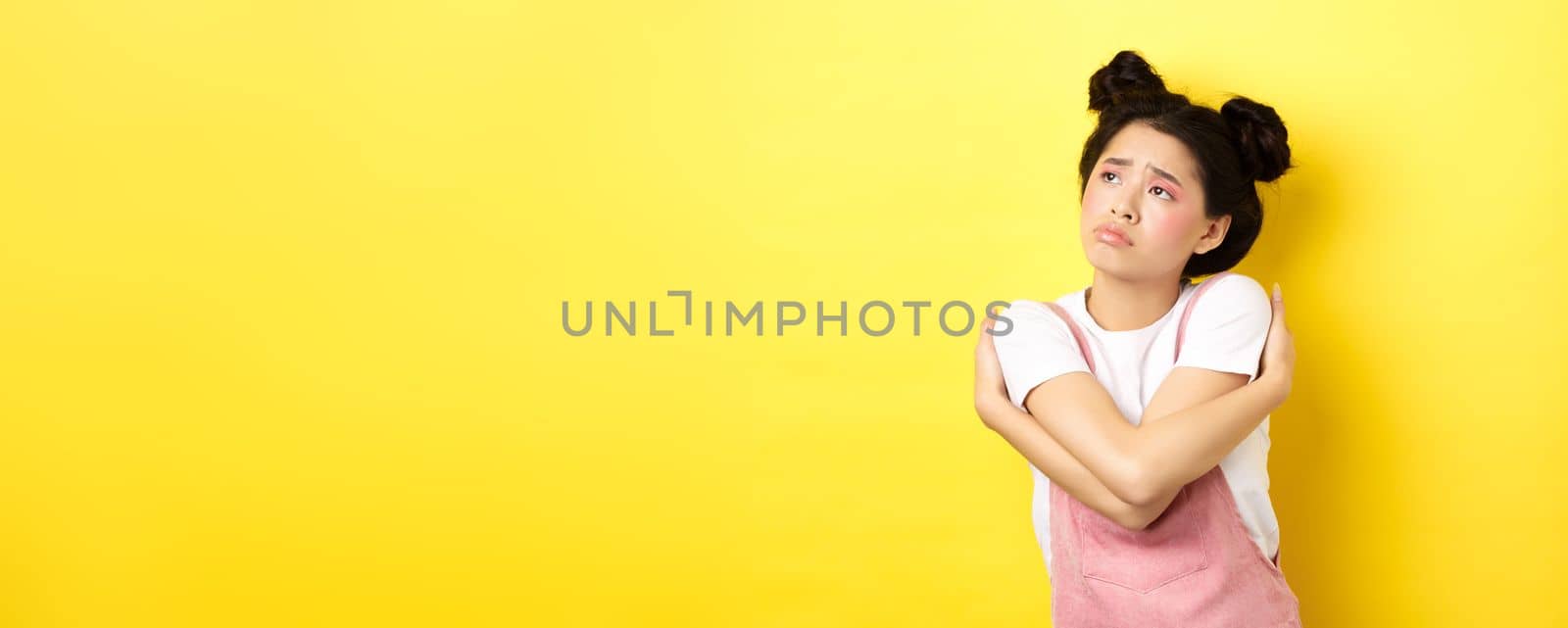 Sad lonely asian girl looking aside with upset and gloomy face, hugging herself, dreaming of love and boyfriend, yellow background by Benzoix