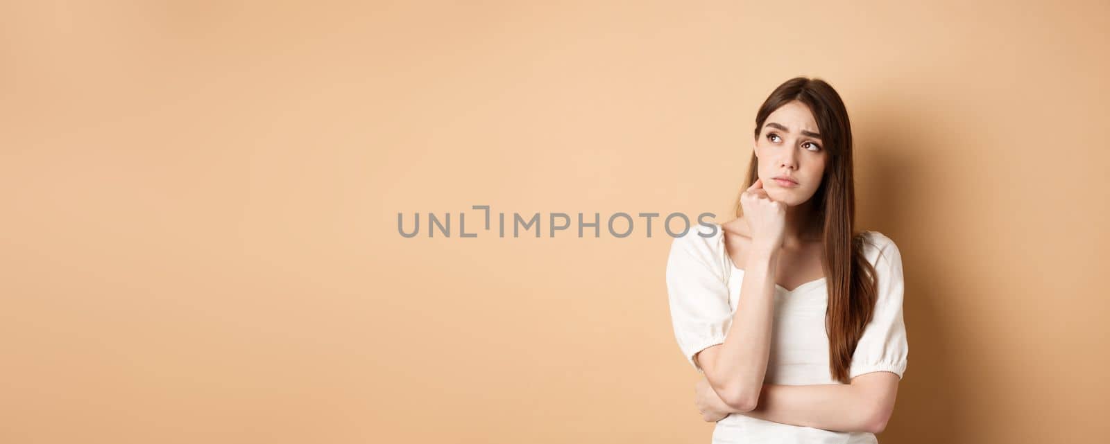 Sad young woman looking pensive at upper left corner, feeling upset or distressed, standing lonely on beige background.