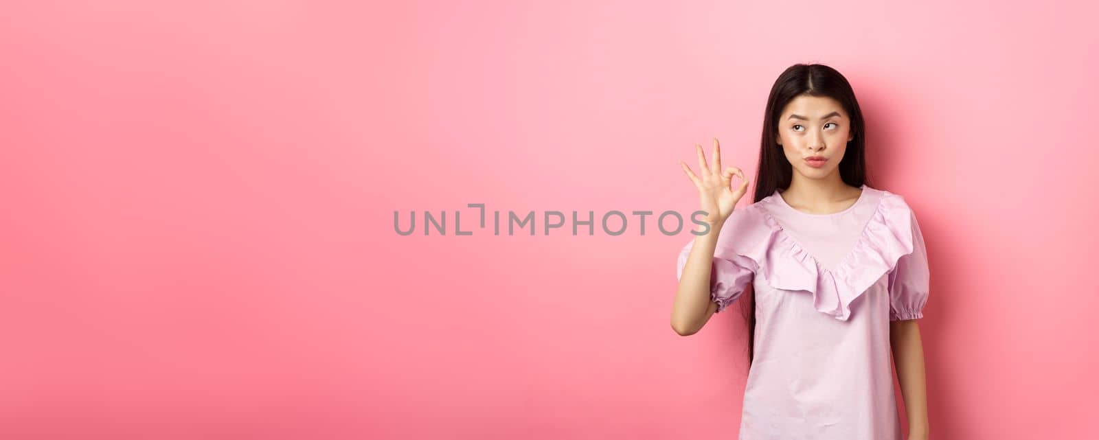 Sassy asian woman in dress show okay sign, looking aside, assure everything good, guarantee all OK, standing pleased on pink background by Benzoix