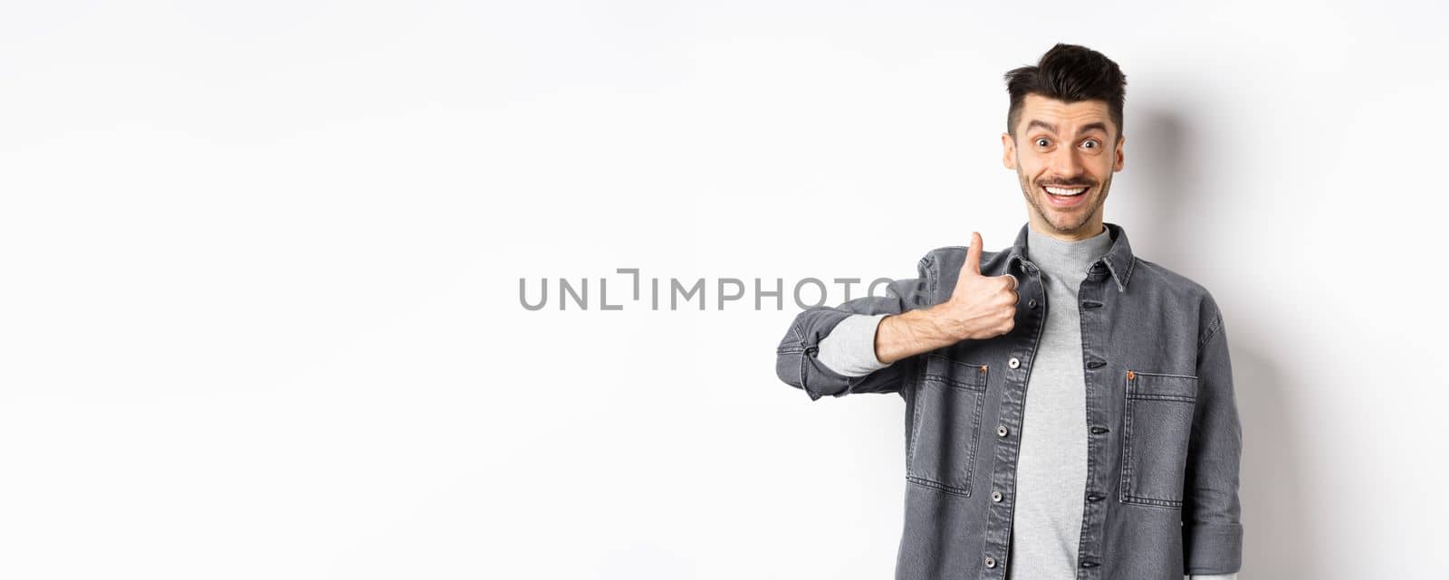 Excited handsome guy showing thumb up and smiling, like good thing, praise awesome choice, looking satisfied and happy, standing on white background by Benzoix