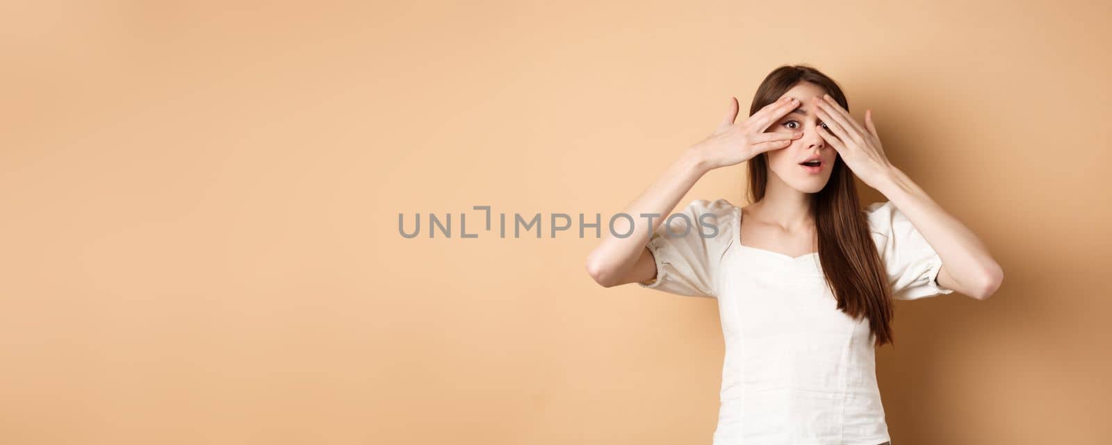 Curious girl peeking through fingers at something interesting, cannot wait to see surprise, look intrigued at camera, beige background.