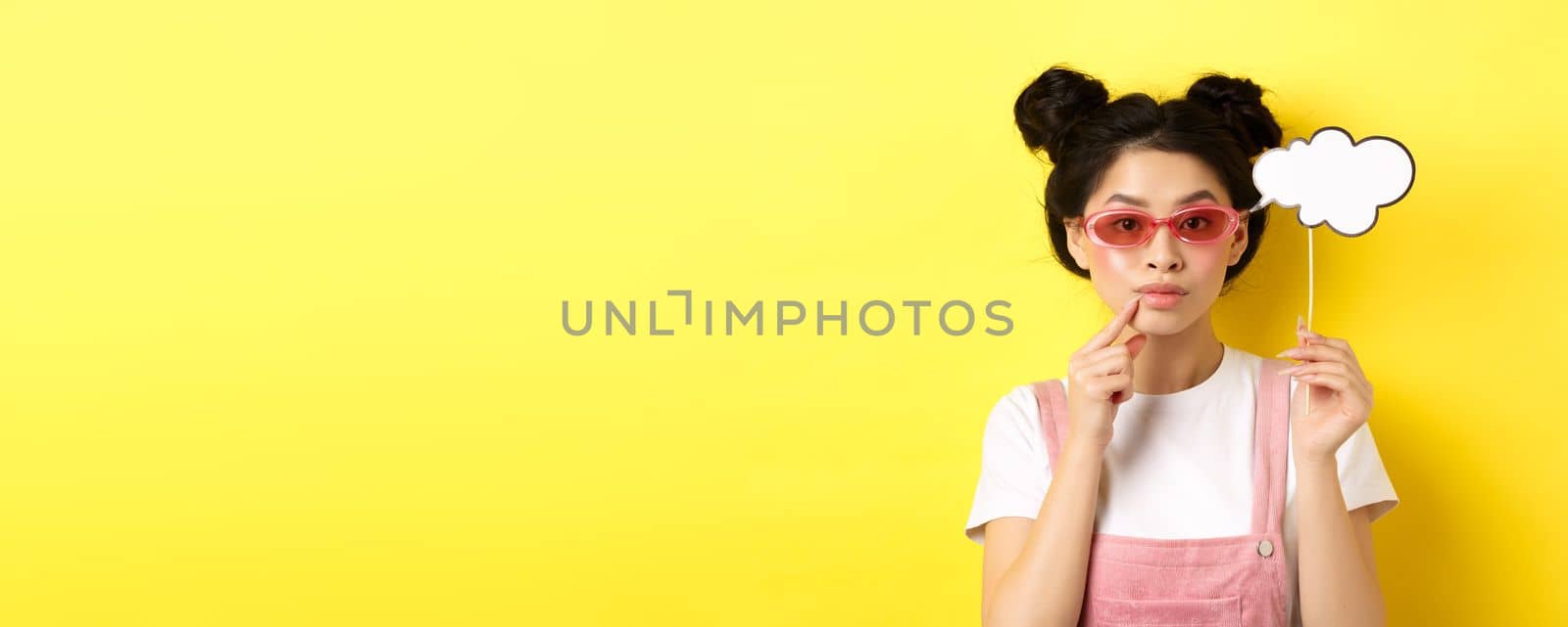 Summer and fashion concept. Stylish asian girl in sunglasses holding comment cloud on stick and looking thoughtful, thinking or saying something, standing on yellow background by Benzoix