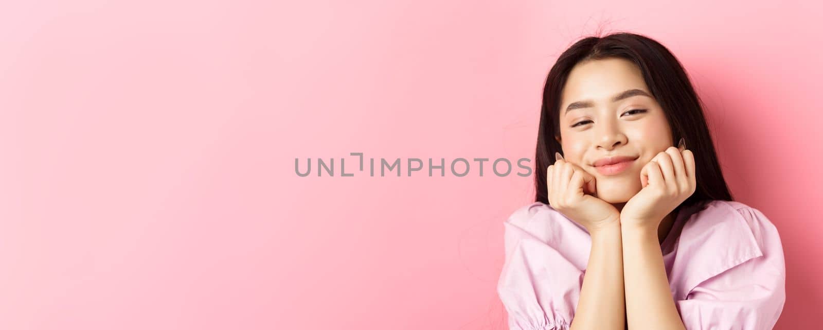 Close-up portrait of dreamy and romantic asian girl, lean face on hands and smiling, looking with admiration and happiness, standing against pink background by Benzoix
