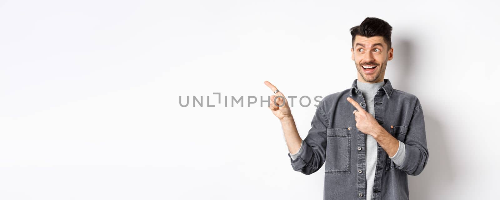 Handsome smiling guy checking out advertisement, looking and pointing left with excited face, standing on white background.