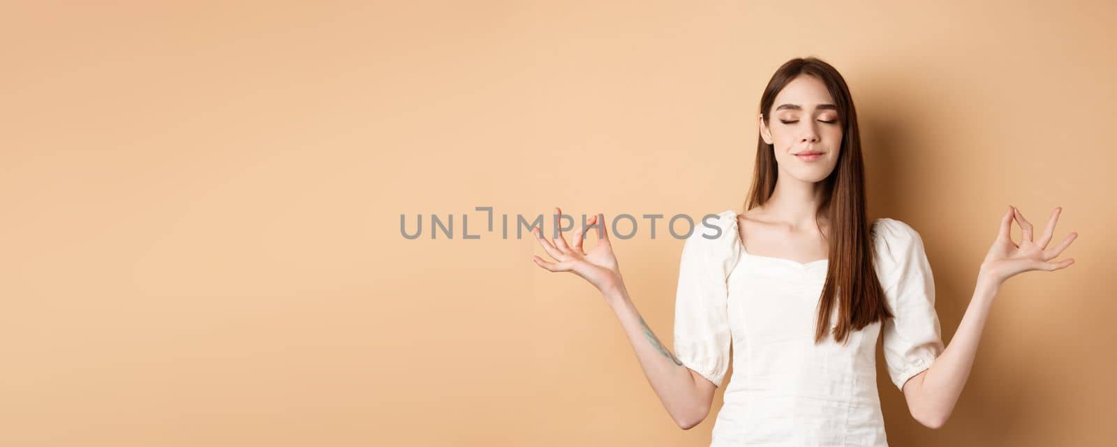 Relaxed young woman meditating, holding hands in yoga zen pose and close eyes, feel peace and smiling, standing on beige background by Benzoix