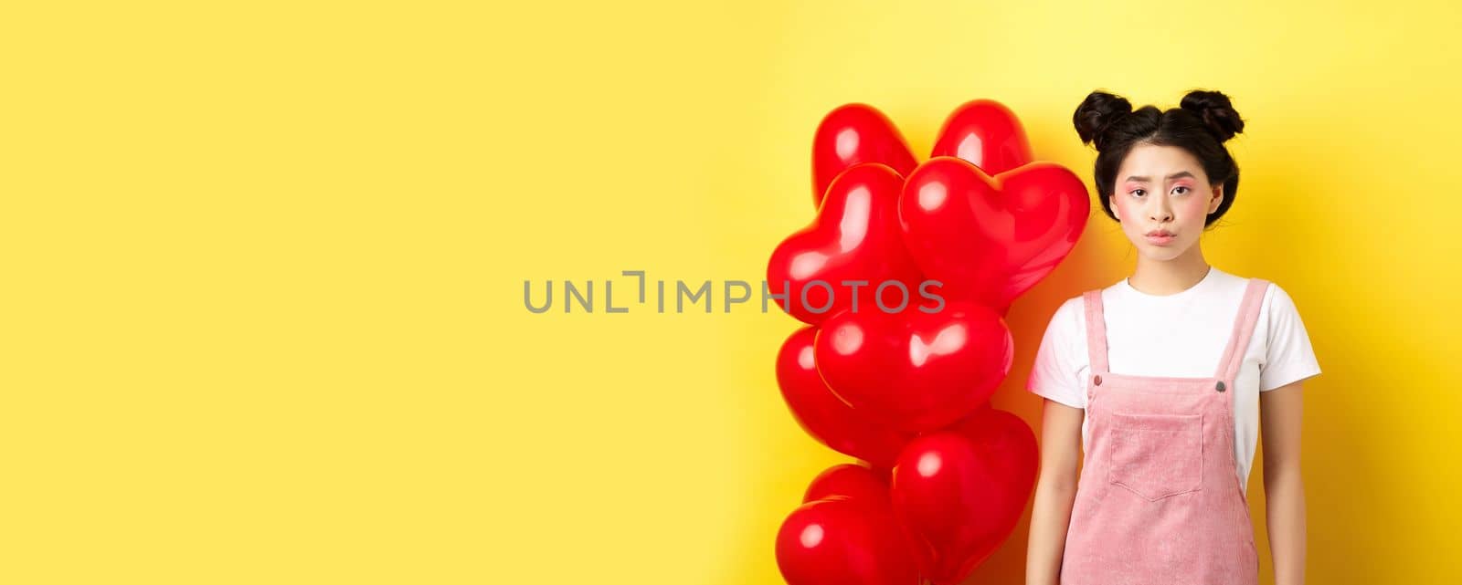 Valentines day concept. Sad and gloomy asian woman in makeup, frowning upset, standing near red hearts balloons and feeling upset, standing on yellow background.