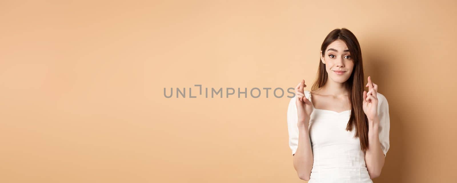 Hopeful tender girl in white blouse making wish, holding fingers crossed and smiling, praying for good luck, waiting for results, beige background.