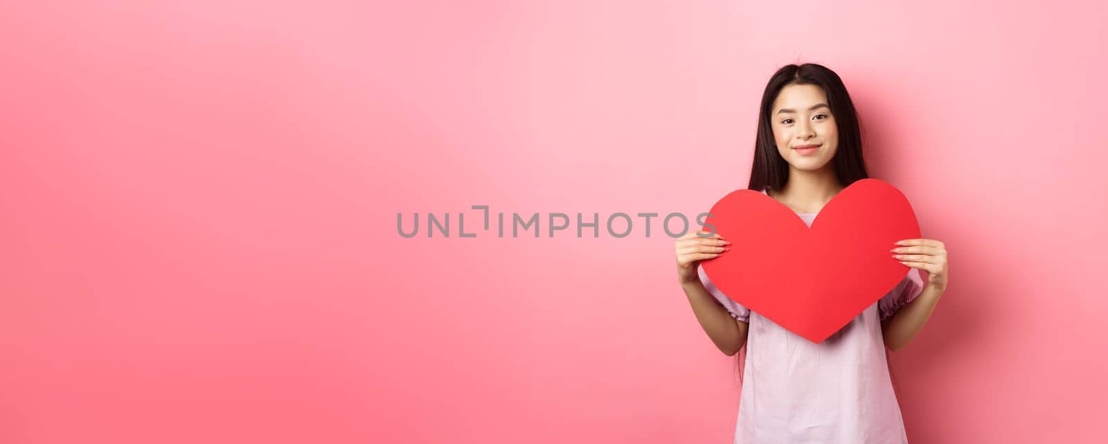 Valentines day concept. Cute teenage asian girl showing big red heart card, falling in love, going on romantic date in dress, smiling tender at camera, pink background.