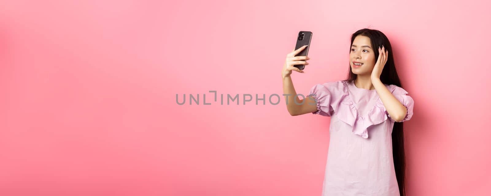 Stylish asian girl blogger taking selfie on mobile phone, posing for smartphone photo, standing in dress against pink background.