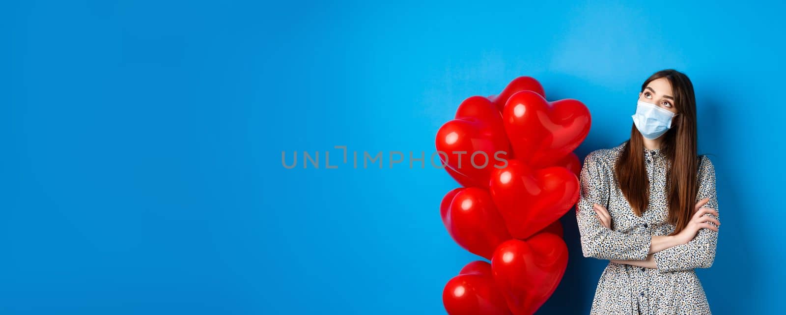 Covid-19, quarantine and health conept. Dreamy beautiful girl in face mask and dress, looking at upper left corner pensive, standing near Valentines day balloons, blue background by Benzoix