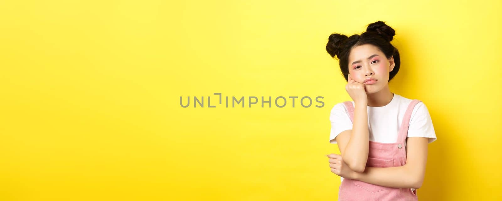Sad and bored teen asian girl standing alone, looking at camera indifferent, staring with boredom, yellow background.