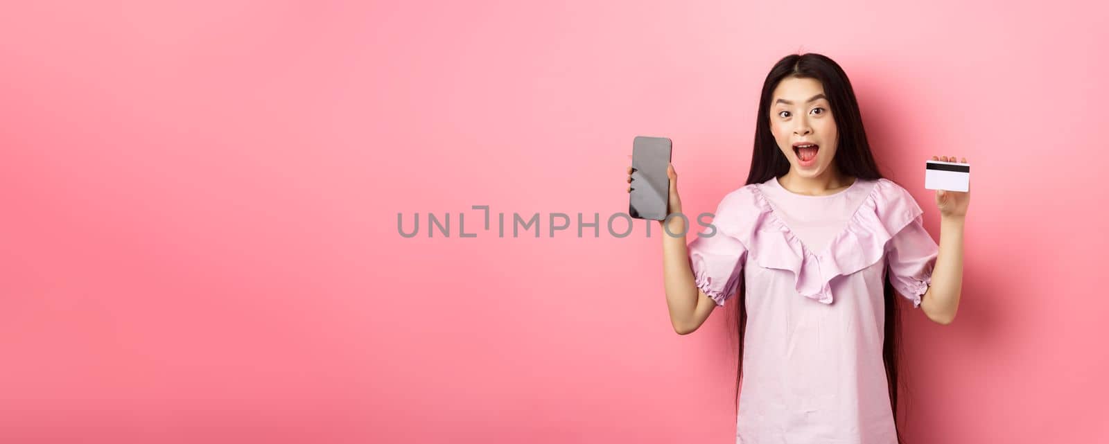 Online shopping. Excited asian woman showing plastic credit card with empty smartphone screen, advertising internet shop, standing on pink background.