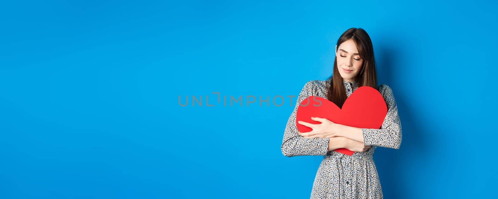 Valentines day. Romantinc girl in dress hugging big red heart cutout, close eyes and smile with dreamy face, imaging sensual date, standing on blue background by Benzoix