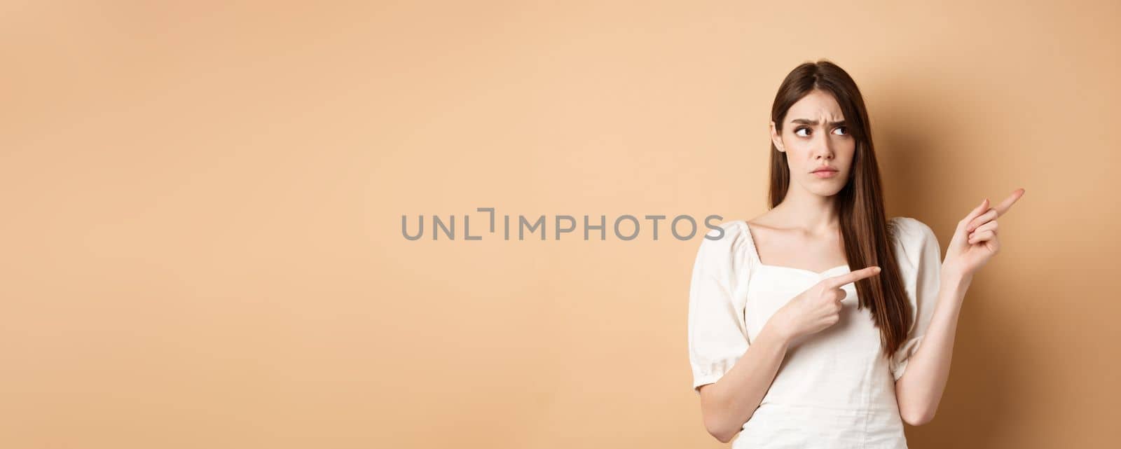 Hesitant young woman look unsure, pointing fingers right while making decision, frowning with puzzled face, standing on beige background by Benzoix