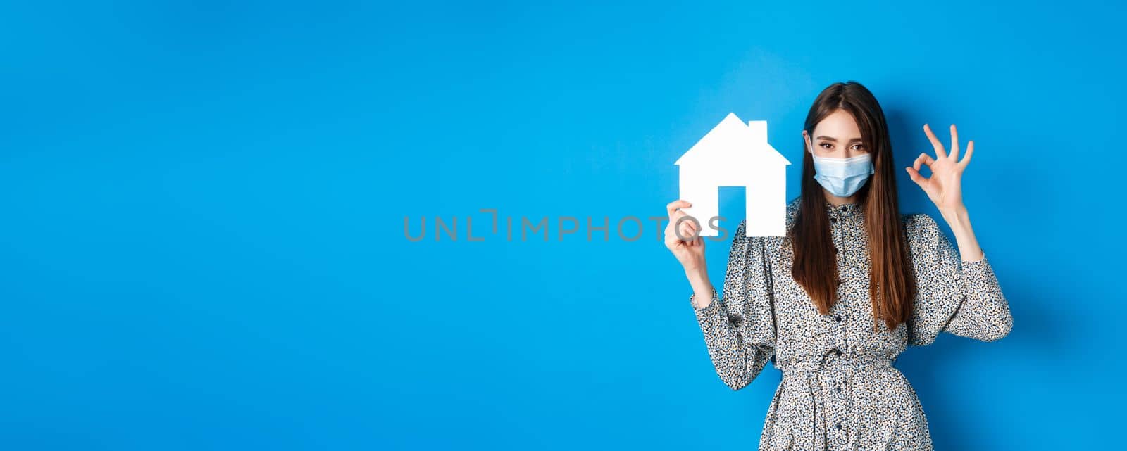 Real estate, covid-19 and pandemic concept. Candid woman in medical mask showing okay sign and paper house cutout, selling property, standing on blue background.