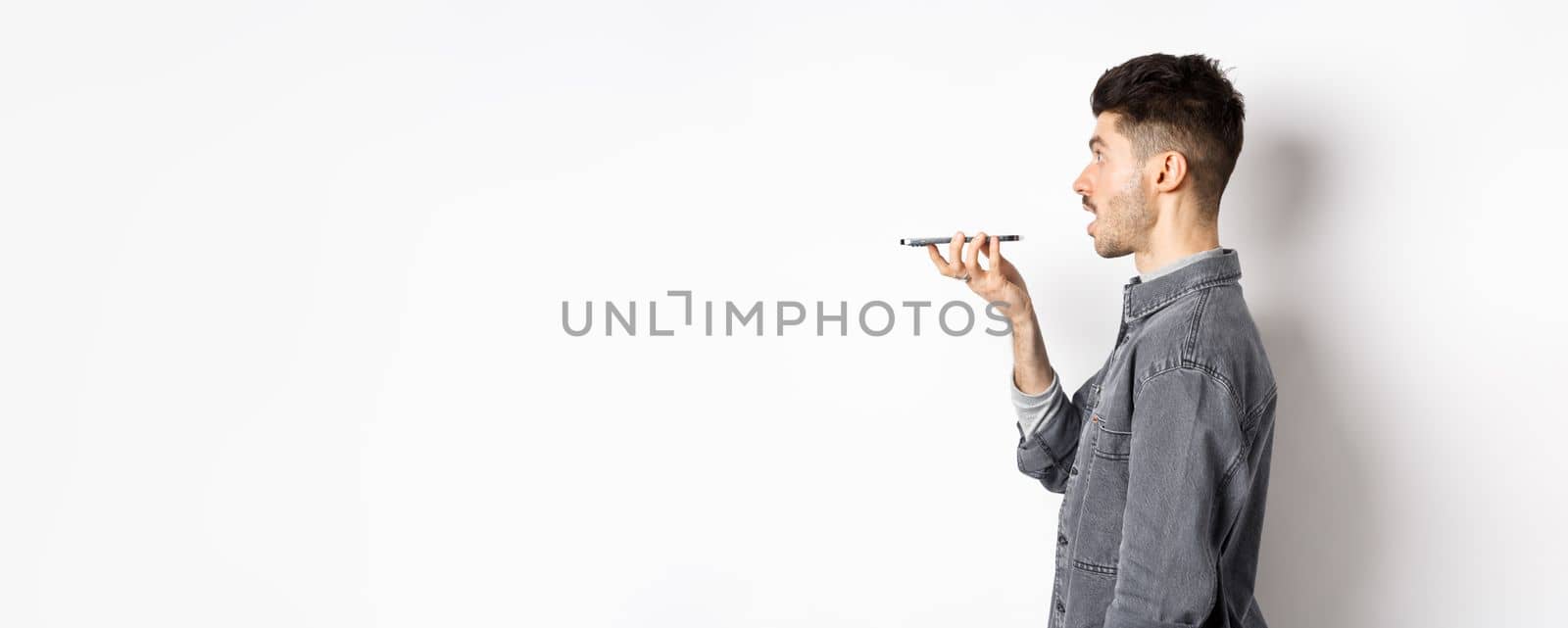 Profile of young man speaking at smartphone, translate with voice-translator app, talking into mobile phone speakerphone, standing on white background.