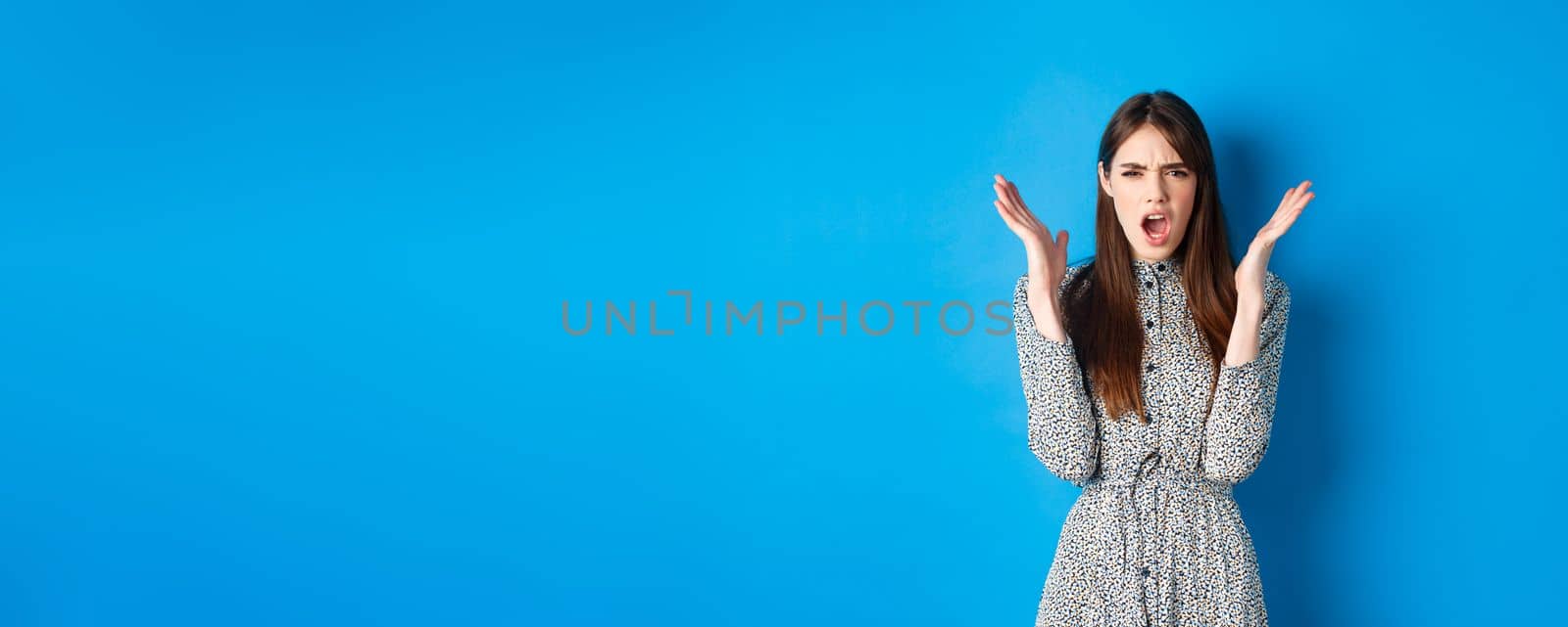 Frustrated and angry woman in dress, scream and frown at camera, raising hands up outraged, having an argument, standing on blue background.