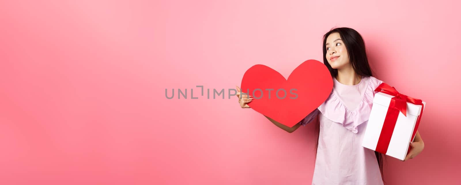 Valentines day concept. Romantic girl holding lover gifts, big red heart card and box with present, looking aside with dreamy look, falling in love, smiling tenderly, pink background by Benzoix